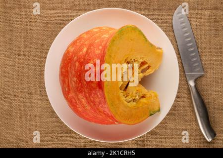 Ein Stück reifen Bio-Kürbis mit weißer Keramikschale auf Jutetuch, Makro, Draufsicht. Stockfoto