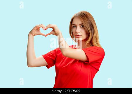 Unterstützendes Frauenporträt. Liebeszeichen. Dankbare zärtliche Dame im roten T-Shirt zeigt auf blauem Hintergrund eine vereinzelte Herzbewegung. Mitgefühl. Versicherungssanc Stockfoto