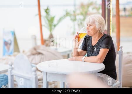 Reife Frau, die in einem Sommercafé an einem Tisch sitzt und Bier trinkt Stockfoto