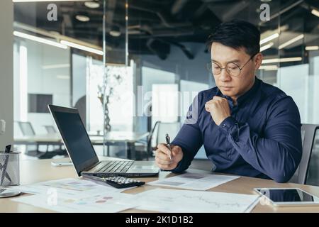 Denkender asiatischer Finanzier in Gläsern Buchhalter, der mit Dokumenten und Konten arbeitet, Laptop benutzt, männlicher Geschäftsmann, der Schreibarbeit beim Sitzen am Schreibtisch im Bürogebäude macht. Stockfoto