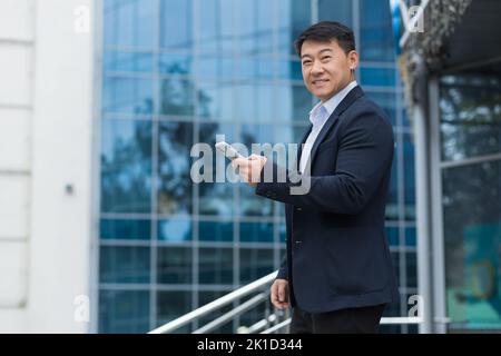 Porträt eines asiatischen Geschäftsmannes vor einem Bürogebäude benutzt ein Mann in einem Geschäftsanzug ein Smartphone, lächelt und schaut auf die Kamera, um die Straße hinunter zu gehen. Stockfoto