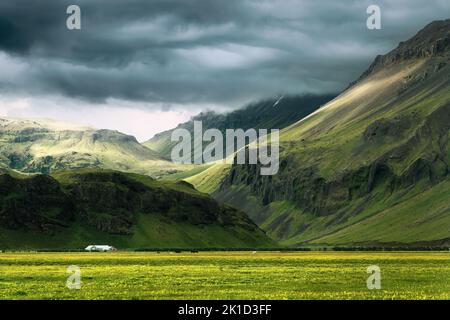 Landschaftlich reizvoller isländischer Berg mit Sonnenlicht, das durch Sturmwolken scheint, und Haus auf dem Feld im Sommer auf Island Stockfoto