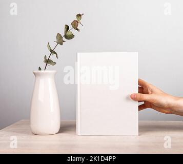 Hand hält Buch Mockup neben Vase mit Blumen an Holztisch. Schönes und zartes Interieur des Literaturliebhabers in minimalistischem Stil. Hochwertige p Stockfoto