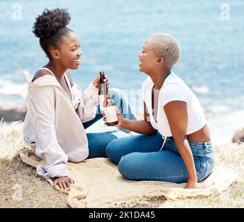 Freunde trinken ein Bier am Strand, während sie sich unterhalten und sich in einem tropischen Sommerurlaub entspannen. Glückliche schwarze Frauen, die Spaß haben, während sie auf einem Meer-Picknick sind Stockfoto