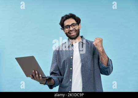 Glücklicher junger arabischer Mann, der den Sieg feiert und seinen Laptop auf Blau isoliert hält. Stockfoto