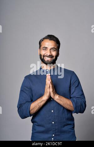 Glücklicher indischer Mann meditiert und hält Hände in namaste isoliert auf grau. Vertikal Stockfoto