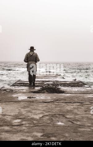 Eine Graustufenaufnahme eines Mantelmannes mit Hut, der an einem rauen Meer unter hellem Himmel steht Stockfoto