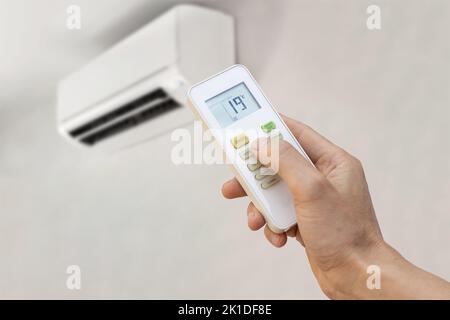 Hand mit Fernbedienung einer Klimaanlage, die die Temperatur auf 19 Grad eingestellt hat. Energiesparkonzept. Stockfoto