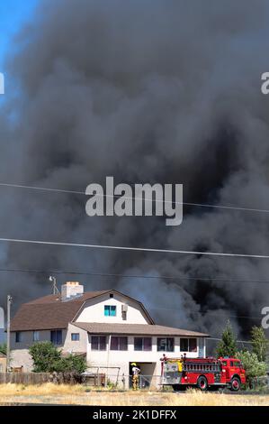 Feuerwehrmann und Feuerwehrauto vor einem Hausbrand mit riesigen Wolken wehenden Rauchs. Stockfoto