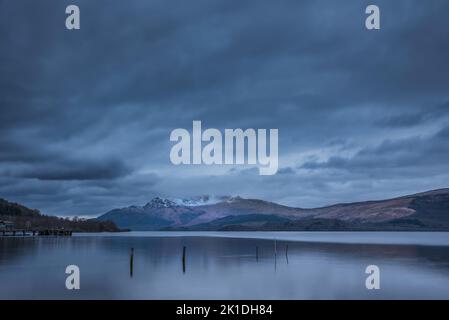 Atemberaubendes Landschaftsbild von Loch Lomond und schneebedeckter Bergkette in der Ferne vom kleinen Dorf Luss aus gesehen Stockfoto