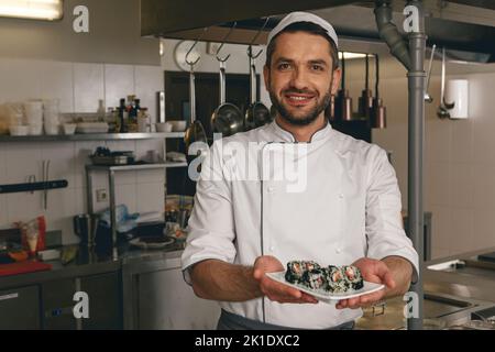 Der gutaussehende Koch des Japanischen Restaurants zeigt einen Teller mit Sushi, der auf der Küche steht Stockfoto