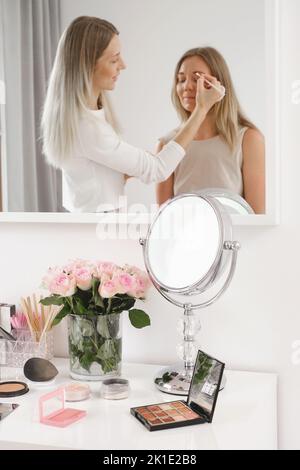 Make-up-Künstler arbeitet mit Modell im Spiegel reflektiert Kosmetik für Make-up und Rosen Blumen auf dem Tisch. Dekorative Kosmetika, Pinsel, Augen Schatten mit Stockfoto