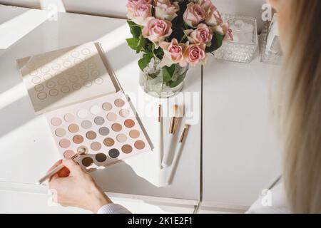 Hand taking Kosmetik für Make-up, Rosen Blumen auf dem Tisch. Dekorative Augenschatten mit Fundament auf Frisiertisch im Schönheitssalon. Auswahl des Künstlers Stockfoto