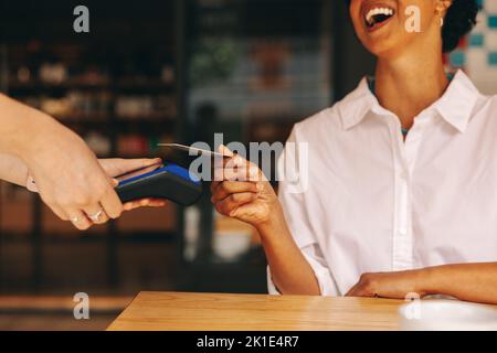 Glückliche Kundin scannt ihre Kreditkarte auf einem Kartenautomaten, um ihre Rechnung in einem Café zu bezahlen. Fröhliche Frau, die eine bargeldlose und kontaktlose Transaktion macht Stockfoto