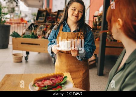 Freundliche Kellnerin mit Down-Syndrom serviert einem Gast ein Sandwich und Kaffee in einem trendigen Café. Professionelle Frau mit einer intellektuellen Behinderung Stockfoto