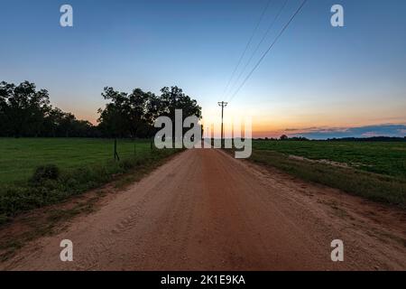 Leere rote Feldstraße, die in den Sonnenuntergang führt, mit Weide auf der einen Seite und Erdnussfeld auf der anderen. Stockfoto