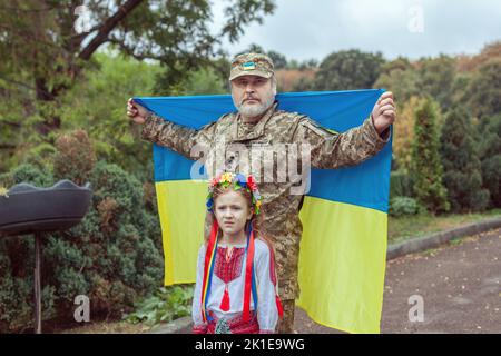 Porträt eines ukrainischen Soldaten mit seiner kleinen Tochter. Sie stehen mit der Flagge der Ukraine. Stockfoto