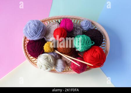 Rot, gelb, weiß, lila, braun, Grüne Fadenkugeln in einem beigen Korb mit Stricknadeln aus Bambus auf einem bunten Hintergrund. Stockfoto