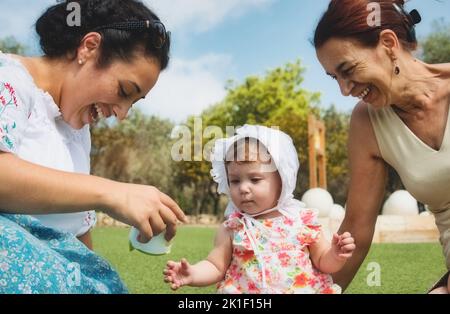 Eine glückliche Familie von 3 Generationen, Baby, Mutter und Großmutter, die auf dem Gras sitzt und im Freien in einem öffentlichen Park mit blauem Himmel spielt Stockfoto