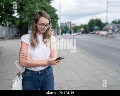 Schöne junge weiße kaukasische Mädchen steht auf dem Bürgersteig und wartet auf die öffentlichen Verkehrsmittel. Langes Blondie-Haar. Fahrzeuge im Hintergrund Stockfoto