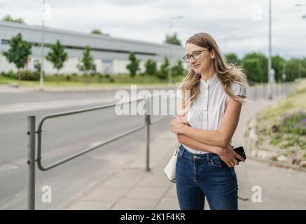 Schöne junge weiße kaukasische Mädchen steht auf dem Bürgersteig und wartet auf die öffentlichen Verkehrsmittel. Langes Blondie-Haar. Fahrzeuge im Hintergrund Stockfoto