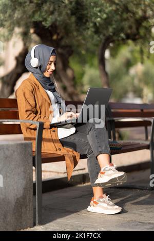 Muslimische Frau, die im Freien einen Laptop und Kopfhörer benutzt Stockfoto