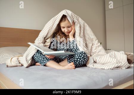 Niedliches kleines Teenager-Mädchen, das auf dem Bett unter der Decke liegt und Buch liest Stockfoto