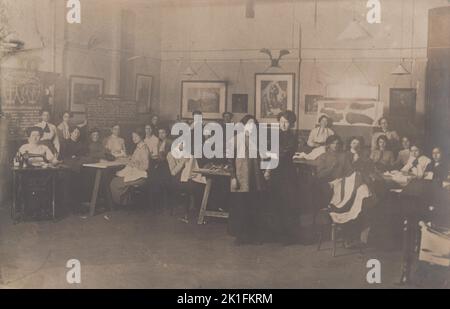 Frauen Schneiderei oder Dressmaking Klasse, Anfang 20. Jahrhundert. Das Foto zeigt junge Frauen, die in einem Klassenzimmer saßen, eine arbeitet an einer Tretnähmaschine und die anderen arbeiten in der Hand. Eine Frau, die in der Mitte des Bildes steht, probiert ein teilweise fertiggefertigtes Kleidungsstück an. Schriftliche Anweisungen und Diagramme werden auf Tafeln aufgestanzt und auf der Rückseite sind Stücke maßgeschneiderter Kleidungsstücke zu sehen Stockfoto