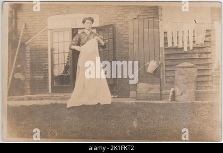 Hängen aus der Wäsche, Anfang des 20.. Jahrhunderts. Informelles Foto einer edwardianischen Frau, die am Waschtag in ihrem Garten steht, mit einem Laken oder einer Decke, die sie kurz vor dem Aufhängen an der Wäscheleine steht. Strümpfe und andere Gegenstände sind bereits zum Trocknen aufgehängt. Im Hintergrund sind auch ein Metallstaubbehälter und eine Gießkanne zu sehen. Stockfoto