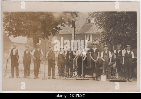 Straßenmender aus dem frühen 20.. Jahrhundert, fotografiert in Arbeitskleidung, mit Spaten und Bürsten. Sie werden vor einer von Pferden gezogenen Dampfmaschine fotografiert Stockfoto