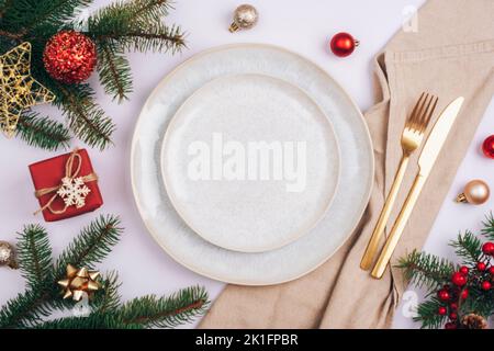 Stilvolle Tischunterlage, Tannenzweige, Sterne und Kugeln mit weißem Teller, Besteck und beige Serviette auf weißem Tisch. Weihnachtskonzept. Draufsicht, flach liegend. Stockfoto