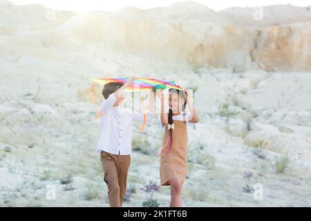 Ein Mädchen und ein Junge tragen einen bunten Drachen über ihren Köpfen, während sie in den Bergen spazieren. Stockfoto