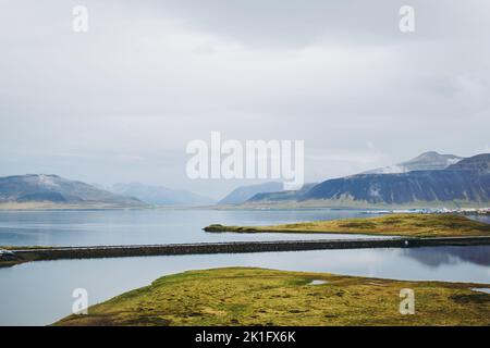 Bunte isländische Landschaften unter grauem, schwerem Himmel. Grünes nasses Gras, schöner See und schneebedeckte Gipfel der Berge spiegeln sich im Wasser des Sees wider. Kirkj Stockfoto