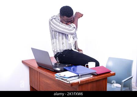 Junger Geschäftsmann, der im Büro auf dem Tisch sitzt, eine optische Brille trägt und im Ellbogen hustet. Stockfoto