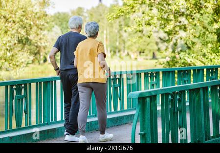 Rückansicht Frau und Mann mittleren Alters tragen aktive Kleidung, spazieren entlang der Brücke im Sommerpark, genießen Sie morgens einen sportlichen Spaziergang zusammen draußen. Gesunde Lif Stockfoto