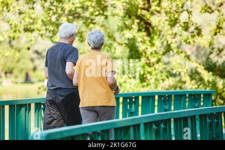 Rückansicht Frau und Mann mittleren Alters tragen aktive Kleidung, spazieren entlang der Brücke im Sommerpark, genießen Sie morgens einen sportlichen Spaziergang zusammen draußen. Gesunde Lif Stockfoto