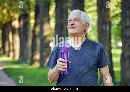 Älterer Mann in Sportbekleidung hält wiederverwendbare Plastikflasche stehen oder joggen im Sommerpark, sich um Gesundheit kümmern, genießen Sie morgens trainieren fühlt sich glücklich. Hea Stockfoto
