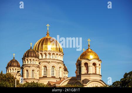 Eine Kirche mit einer gelben Mauer und einer spitzen goldenen Spitze mitten im Stadtzentrum mit mehreren Fenstern und Bäumen daneben Stockfoto