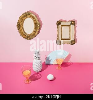 Tropischer Hintergrund mit weißer Ananas und Zitrone, Trinkgläsern und blauem Papierventilator auf rosafarbenem Tisch und goldenen Vitagebilderrahmen auf rosafarbener Wand. Stockfoto