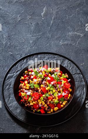 Schwarzer Bohnensalat mit schwarzäugigen Erbsen, Pfeffer, roten Zwiebeln, Mais, Kirschtomaten und Koriander in schwarzer Schüssel, vertikale Ansicht, Nahaufnahme Stockfoto