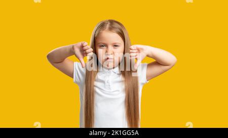 Frech Kind. Geste nicht gefallen. Ablehnungssignal. Porträt eines unzufriedenen, skeptischen kleinen Mädchens, das den Daumen nach unten zeigt, Missbilligungsschild isoliert auf gelbem c Stockfoto