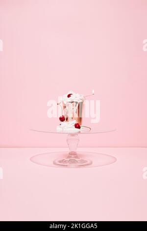 Goldenes Papier mit schmelzender Schlagsahne, Kirschen und brennender Kerze auf Glaskuchen stehen vor rosa Hintergrund. Cupcake im Sommer. Minimalkonzept. Stockfoto