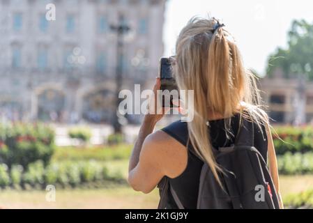 Rückansicht einer blonden Frau, die ein Smartphone-Foto des Buckingham Palace aus dem Fokus nimmt. Stockfoto