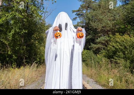 Witziges Halloween. Ein kleines Kind in einem weißen Anzug, das Körbe in Form eines Kürbisses, einen gruseligen Halloween-Geist, auf der Straße hält. Ein Kind, das als Hal gekleidet ist Stockfoto