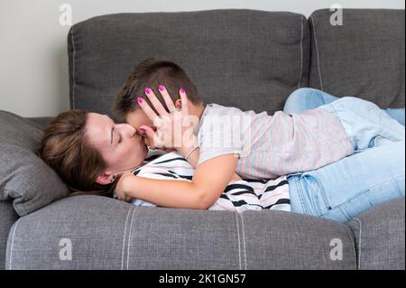 Frau liegt auf dem Sofa und küsst ihren kleinen Sohn mit der Wange. Mama mit kleinen Jungen freudig verbringen Zeit in gemütlichen Wohnzimmer zu Hause. Stockfoto