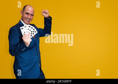 Ein kahlköpfiger Kaukasusmann, der in einem blauen Anzug, einem weißen Hemd und einer orangefarbenen Krawatte gekleidet ist, hält fünf Spielkarten, die in einem Pokerspiel einen geraden Kleeblätter bilden. Stockfoto