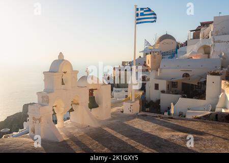 Oia, Santorini - September 11 2022: Das goldene Abendlicht wirft lange Schatten von den traditionellen griechisch-orthodoxen Kirchenglocken neben einer griechischen Flagge Stockfoto