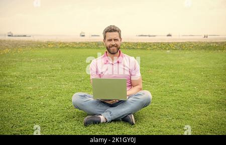 Glücklicher Geschäftsmann mit Laptop zum Bloggen auf grünem Gras, Freiberufler Stockfoto