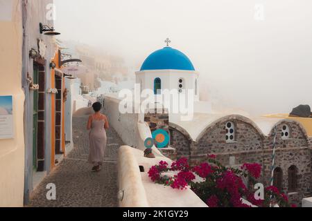 Oia, Griechenland - 9. September 2022: Eine Frau geht entlang einer malerischen Kopfsteinpflasterstraße entlang der traditionellen, blauen Kuppel der griechisch-orthodoxen Kirche Panagia Agion Stockfoto