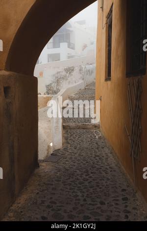 Ruhiger, enger, gepflasterter Straßenweg an einem nebligen, nebligen Morgen im alten Stadtdorf Oia auf der griechischen Insel Santorini, Griechenland. Stockfoto